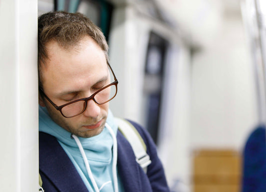 Depicted is a masculine person with glasses leaning against a wall; he appears to be asleep. the rest of the image is blurry to focus on the man. Chronic Fatigue