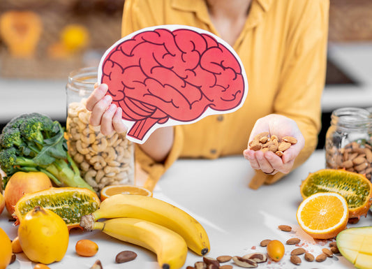 The person is holding a paper cut out of a pink brain in one hand and in the other a handful of almonds. Around here on a table are various nuts, fruit, and vegetables. broccoli, banana, clementines, oranges, almonds, cashews, apples. horned melon and Lemons Gut Brain