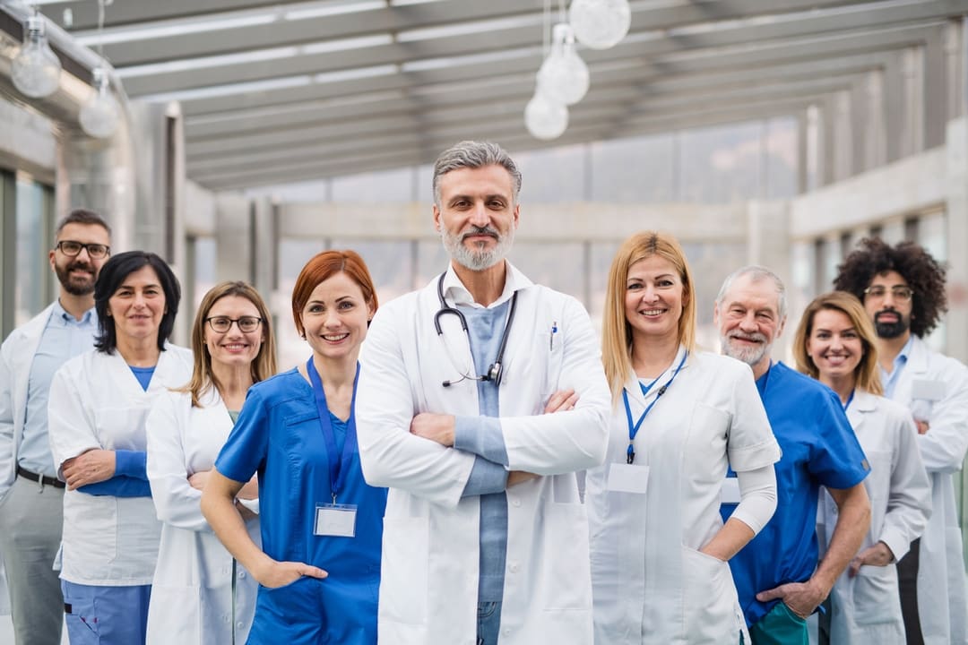 A picture of nine doctors standing with their hands either in their pockets or crossed. They are wearing a mix of white and blue.