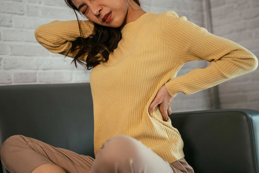 woman with black hair and yellow sweater sitting cross legged on a green couch holding her neck and back in pain Chronic Pain