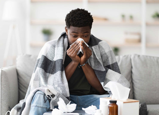 Man sitting on a gray couch with a gay plaid blanket over his shoulders as he blows his nose. in the foreground is a box of tissues and several crumples use tissues, along with some medison Immune System Problems
