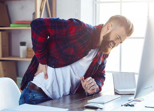 Picture of a man leaning forward at this computer desk while holding his side/back. His face is scrunched in pain. Back Pain