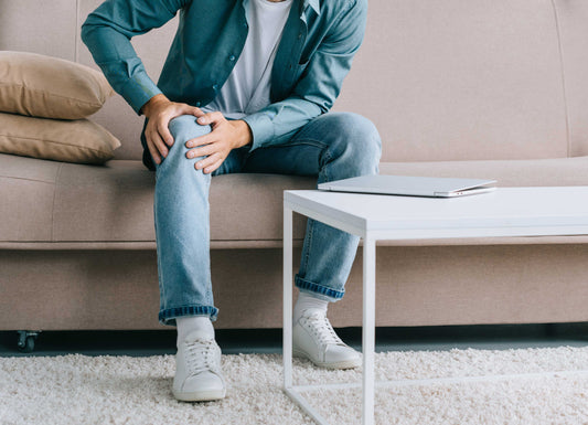 cropped picture of a person sitting on a couch holding their knee in pain. Knee Pain