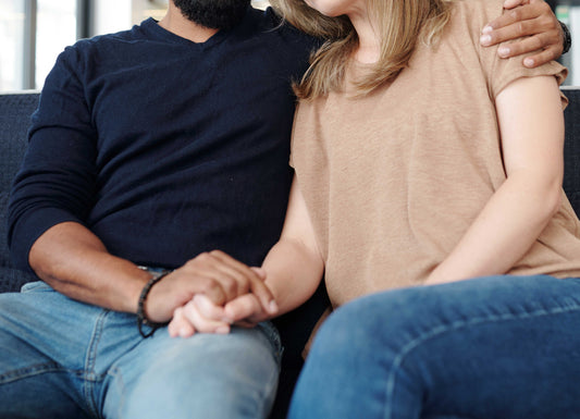 A couple sitting together. The man has an arm around the woman and they are holding hands in comfort. Miscarriage