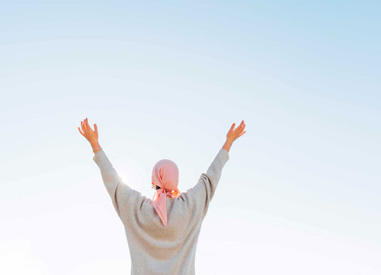 Depicted is a feminine person reaching up to the light blue sky, their back facing us. the person is wearing a gray sweater and has their hair covered in a pink scarf Cancer