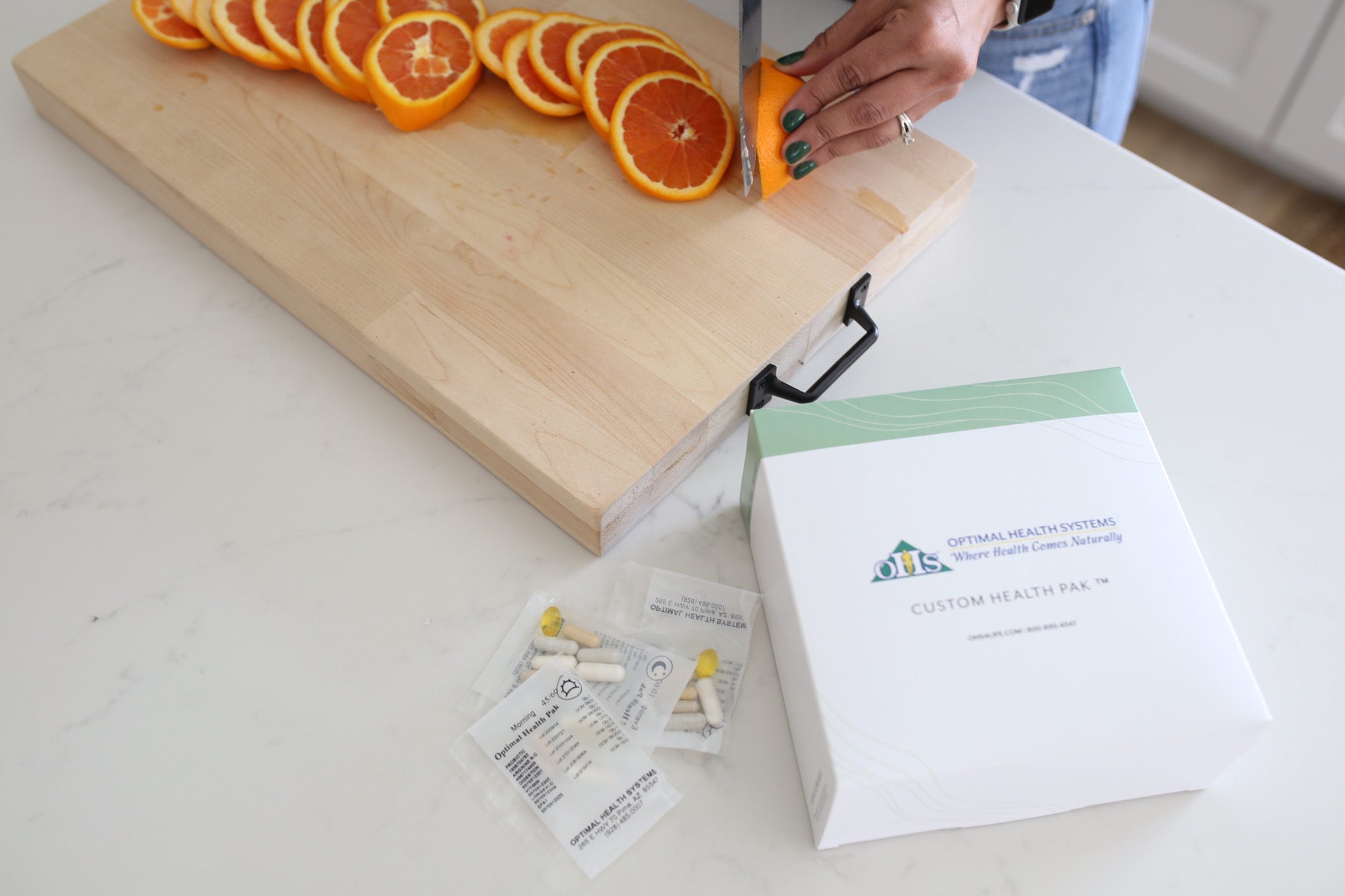A zoomed-in picture of a countertop that someone is using to cut oranges on a wooden cutting board. There's an Optimal Custom Healthpak with 3 pak packets sitting next to the cutting board.