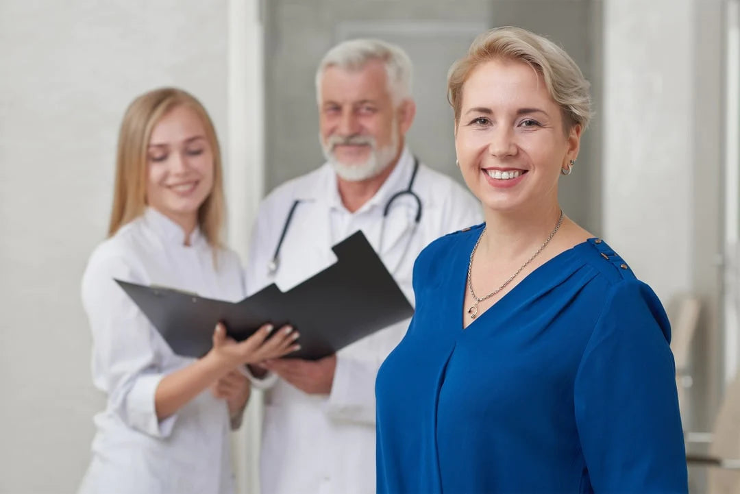 In the foreground is a happy patient wearing blue, and in the background are two happy doctors looking at a clipboard.